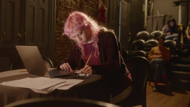 Young female playwright with pink hair using laptop, writing notes and posing for camera with smile on theater stage. Medium long shot, video portrait