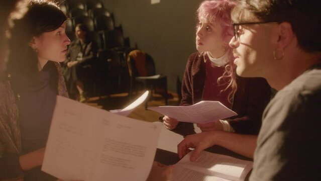 Group of young drama students sitting around the table on theater stage, reading play scripts and discussing their roles during rehearsal of performance