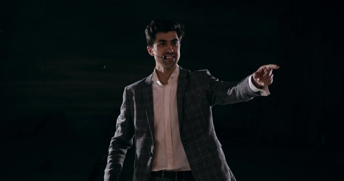 Showman performs on stage in dark black background Young male entertainer, presenter or actor on stage pointing to side Public speaker speaking at the microphone, you can do everything, never give up