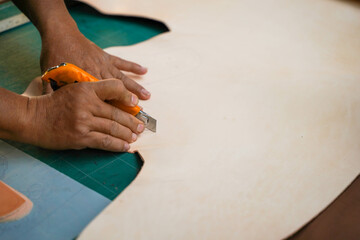 Closeup and crop hands of leather craftsman is using a cutter knife to cut a piece of white leather large sheet according to the design to bring out to sew into products for customers. - Powered by Adobe