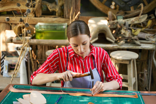 Closeup young female leather maker using a small hummer with leather punching steel to make a leather belt for customers.