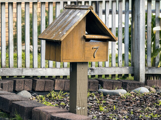 Stand along wooden brown post mounted traditional gable roof letterbox with street number 7. Square mounting post. Newspaper slot above main mail slot.