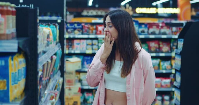 dark-haired customer examines goods on counter in supermarket is outraged by overpriced. young woman is surprised by overpriced goods on background of the store. Lady at loss from the inflated cost