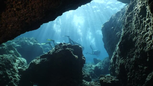  scuba divers exploring around a reef underwater deep blue water big rocks and bubbles ocean scenery 