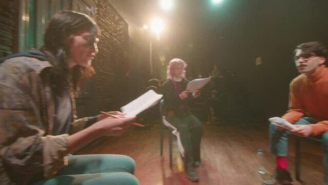 Young actors sitting in a circle on stage, reading play scripts and practicing speech during rehearsal in a theater or drama class. PAN handheld shot