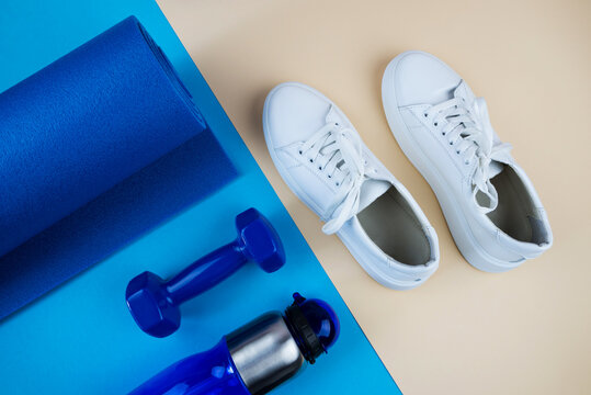 The concept of sports accessories. A photo of white sneakers, blue dumbbells, a blue exercise mat and other sports equipment against a pastel beige background.