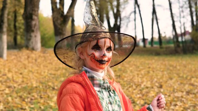 A child girl in an orange jacket, a witch's hat and with makeup on her face holds a decoration of skeletons in her hands. Halloween celebration. Horizontal video