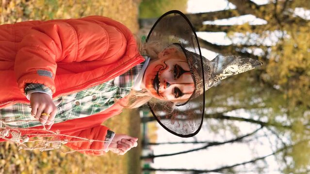 A child girl in an orange jacket, a witch's hat and with orange makeup on her face holds a decoration of skeletons in her hands. Halloween celebration. Vertical video