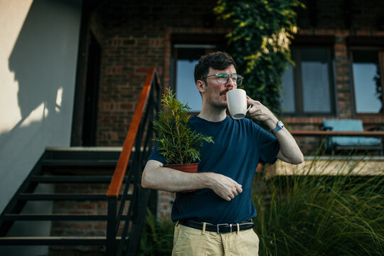 Shot of a man standing in front of his house, holding a plant and drinking a coffee.