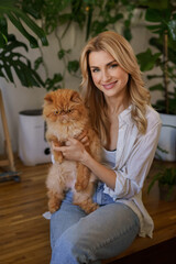 A charming blonde girl in a white shirt and denim jeans cuddles her ginger cat while sitting on a table surrounded by potted plants