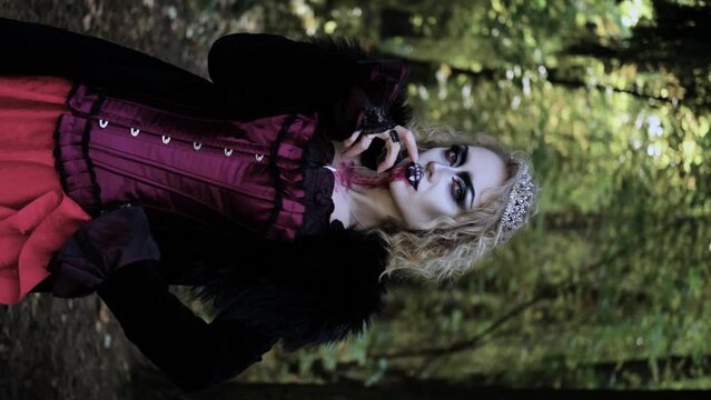 A woman in the image of a vampire or witch with makeup and medieval clothes poses for the camera in the forest. Vertical video