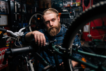 Portrait of bearded technician male fixing mountain bicycle standing on bike rack working in bike repair shop with dark interior, looking at camera. Concept of maintenance of bicycle transport.