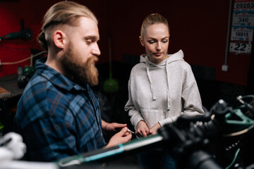 Fototapeta premium Close-up of confident cycling repairman with beard communicating with attractive blonde female client, talking problem of bicycle, detected during diagnostics in repair shop with dark interior.
