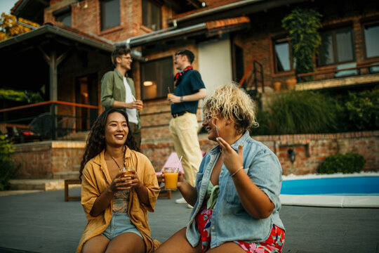 Two diverse female friends sitting and drinking cocktails, men standing and talking in the background during a house pool party.