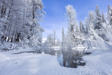 Colorful landscape with snowy trees, river with reflection in water in cold Snow covered trees, lake, sun and blue sky. Beautiful forest in snowy winter
