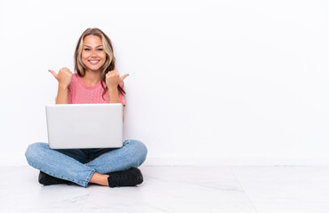 Young Russian girl with a laptop sitting on the floor isolated on white background with thumbs up gesture and smiling