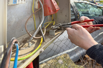 An electrician connects wires in a switchboard outdoors. The hands of an electrician close-up with pliers tightens the screw for fastening the ground wire in the shield.