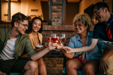 Group of smiling friends sitting and cheering with wine glasses together during a evening on the house porch.