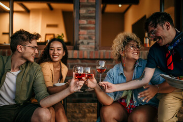 Group of smiling friends sitting and cheering with wine glasses together during a evening on the house porch.