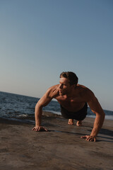 Fototapeta na wymiar Handsome young shirtless man doing push-ups while exercising outdoors with the sea on background