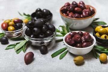 A set of green, red and black olives on a light marble background. Various types of olives in bowls and fresh olive leaves. Vegan. Olive fruits. Place for text. Copy space.