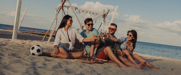 Group of happy young people toasting with beer while enjoying pizza on the beach together