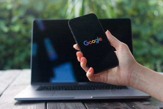 Google logo, American multinational technology company founded by Larry Page, displayed on an iPhone 12 screen with a MacBook Pro in the background.