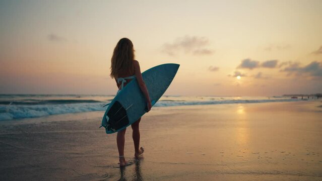 Tourist woman surfer in swimsuit with surfboard walks on ocean sandy beach at sunset choosing spot for surfing. Summer sport activity on tropical resort. Sporty girl. Extreme water sport on vacation.