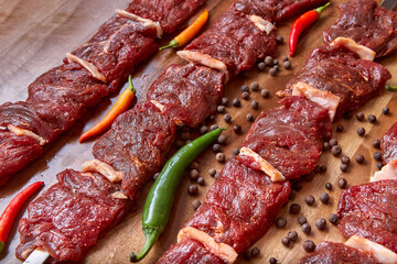 Metal skewers with raw uncooked beef meat for frying on the wooden board with chili peppers and pepper near it, close-up perspective view shallow depth of field. Meat, peppercorns and pepper in focus