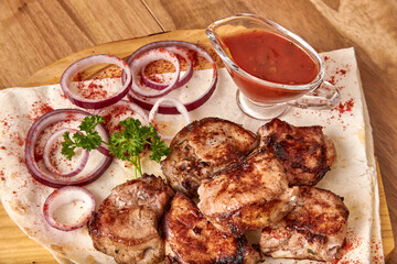 Part of the wooden board with fried beef meat with red onion rings, sauce and pita bread on the wooden table, close-up perspective view, shallow depth of field. Meat and sauce in focus