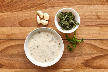 White plate with creamy soup on the wooden table with greens and garlic near it. Top view, flat lay