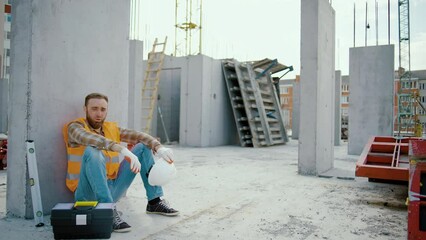 Young tired man worker sitting and takes off white hardhat at construction workplace