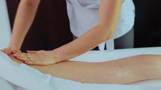 Shooting of young relaxed woman lying on spa massage table in calm spa center room. Hands of female masseuse massaging leg in cozy calm atmosphere indoors. Daytime