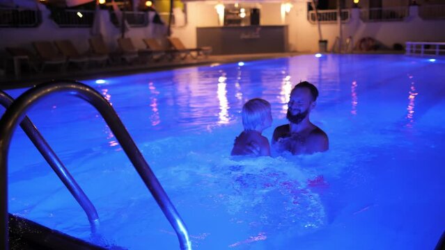 Happy family baby boy and father playing at night illuminated swimming pool luxury resort hotel