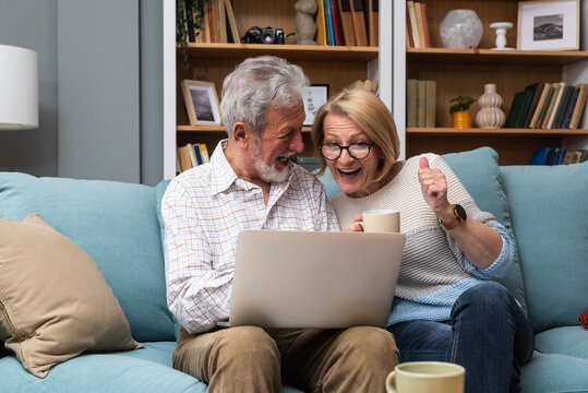 Elderly Retired Couple Sitting At Home Surfing The Internet On A Laptop Looking To Buy A Camper And Sell Their Home To Travel The World. Senior Man And Woman Using Computer To Find Travel Destinations