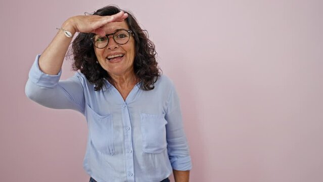 Middle age hispanic woman looking for someone saying hello with hand over isolated pink background
