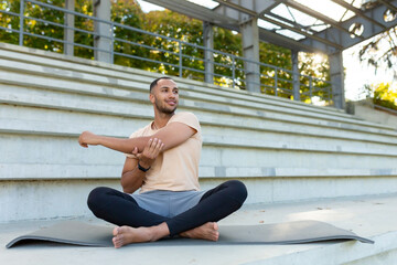 Fototapeta na wymiar A young African American man is playing sports at the stadium. Sits on a mat, performs an exercise, stretches, does yoga