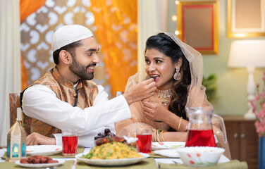 Happy smiling indian muslim husband feeding dates to wife during ramadan dining feast festival celebration at home - concept of relationship, traditional culture and iftar