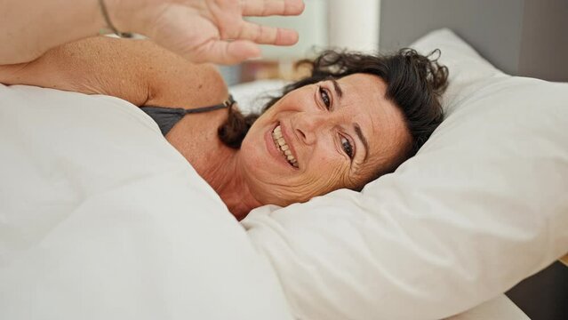 Middle age hispanic woman smiling confident lying on bed saying hello with hand at bedroom