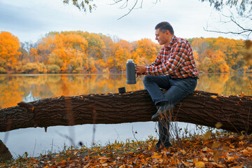 a man sits on a log on the river bank and drinks tea or coffee from a thermos, enjoys the...