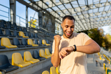 Happy and successful man satisfied with the results of physical training celebrating good results of fitness and running looking at smart watch fitness bracelet walking in the air in the stadium