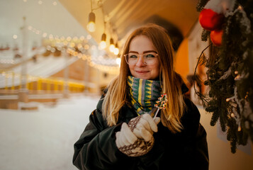 A portrait of a young and smiling woman holding a Christmas tree-shaped lollipop in her hands, standing at a winter fair. The spirit of Christmas, holiday festivities, joy, and sweet treats. - Powered by Adobe