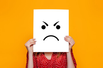 angry emoji. Girl holds a yellow paper poster with emoticon isolated over orange background