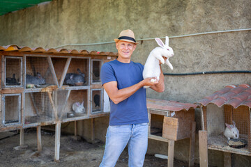 A male farmer holds a white rabbit in his hands on an open farm.