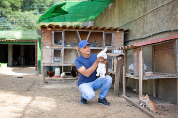 A male farmer holds a white rabbit in his hands on an open farm.