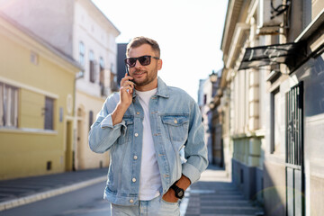 Young man in denim clothes and sunglasses walking down the street and talking on the phone