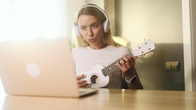 Portrait of pretty young blond woman watch video tutorial on ukulele guitar playing or having personal lesson with teacher distance remote on laptop computer at home Online musical education concept 