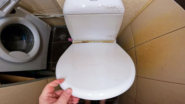 Close-up very dirty toilet bowl with traces of limescale and rust. A man opens the lid of a dirty toilet bowl. Dirty tiled bathroom with washing machine and toilet. Dirty tiles on the floor and walls