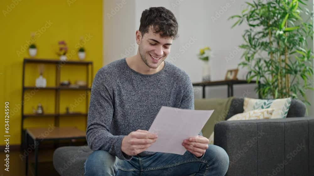 Wall mural Young hispanic man reading document sitting on sofa at home