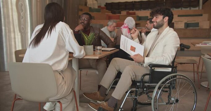 Diverse multiracial business people speaking in cafe bar and being inclusive for disabled colleague on wheelchair while working at the lounge area in the office, trucking